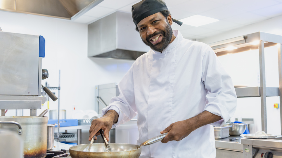 A chef wearing chef whites smiles whilst cooking at the hob with a frying pan
