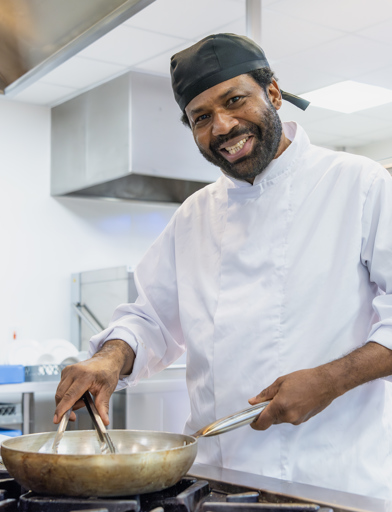 A chef wearing chef whites smiles whilst cooking at the hob with a frying pan