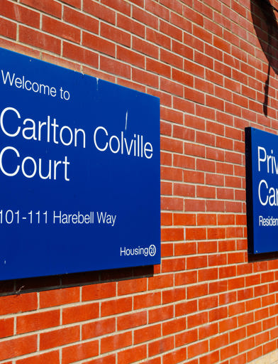 Exterior signage on a red brick wall with a blue sign welcoming visitors to Carlton Colville Court. 