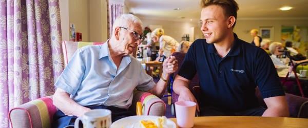 A care worker sitting with a resident in a communal lounge next to a table with cups and a plate of cake on top