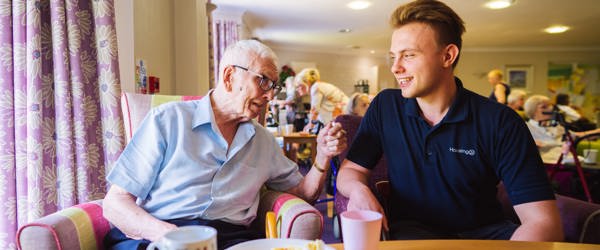 A care worker sitting with a resident in a communal lounge next to a table with cups and a plate of cake on top