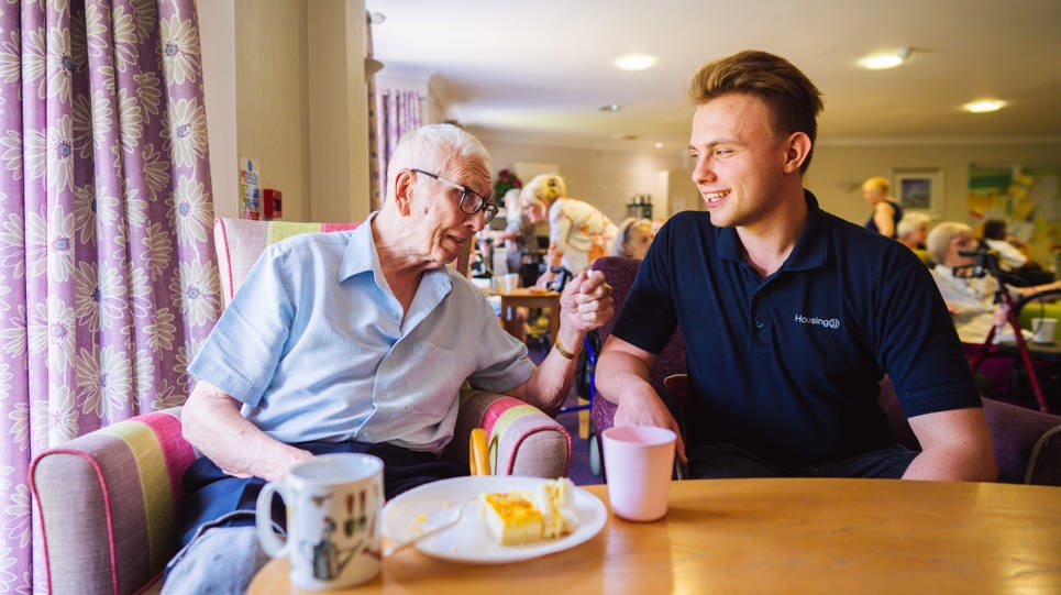 A care worker sitting with a resident in a communal lounge next to a table with cups and a plate of cake on top
