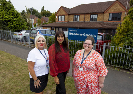Two Housing 21 employees with their lanyards on, smile and pose with MP Sureena Brackenridge outside the blue welcome sign for Broadway Gardens
