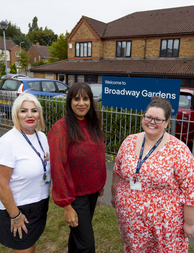 Two Housing 21 employees with their lanyards on, smile and pose with MP Sureena Brackenridge outside the blue welcome sign for Broadway Gardens 
