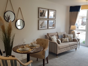 Inside a furnished show apartment at a retirement living scheme. Featuring a dining table with chairs in an open plan living room with a beige sofa on grey carpeted floor