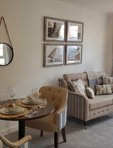Inside a furnished show apartment at a retirement living scheme. Featuring a dining table with chairs in an open plan living room with a beige sofa on grey carpeted floor 