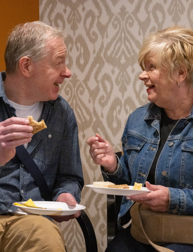 A couple eating sandwiches in a communal lounge while smiling and laughing 