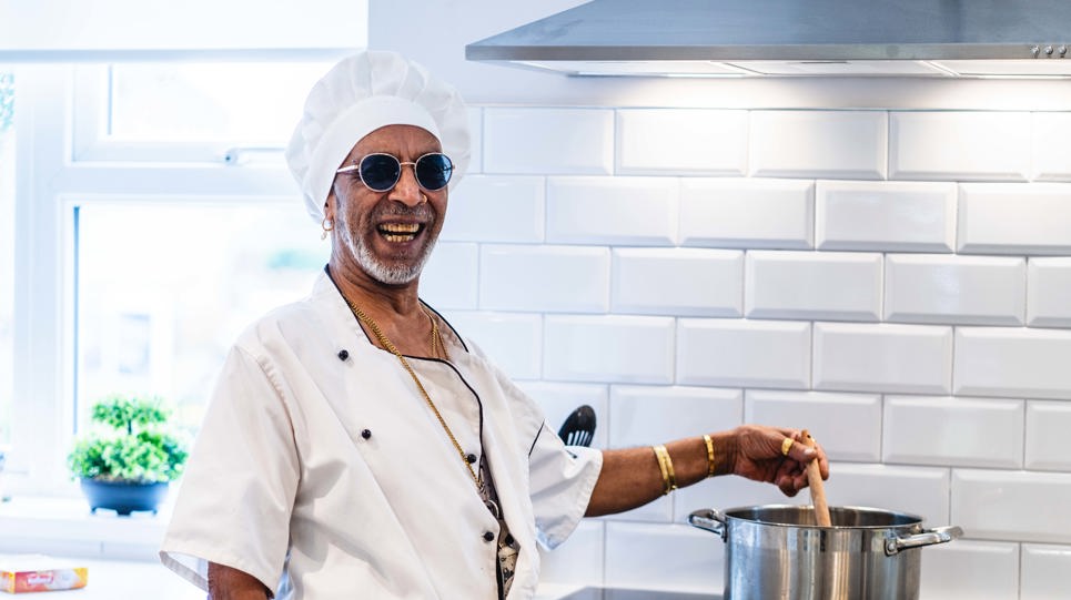 A resident dressed in chef's whites stirs a pot on a cooker and laughs