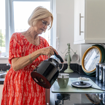 Resident in a red dress pouring a kettle of hot water into a cup in the kitchen of their apartment