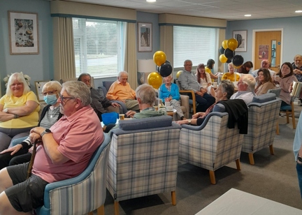 Residents smiling in armchairs in a communal area filled with black and gold balloons.