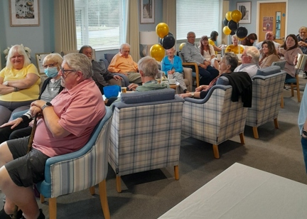 Residents smiling in armchairs in a communal area filled with black and gold balloons.