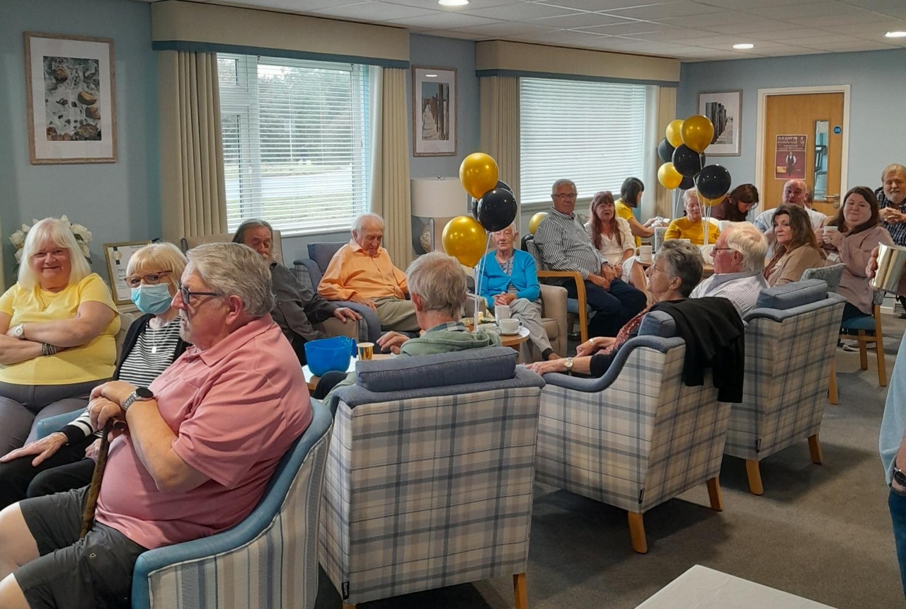 Residents smiling in armchairs in a communal area filled with black and gold balloons.