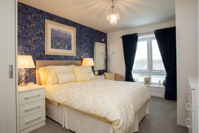 Bright and spacious bedroom featuring a double bed, bedside tables, decorative lighting, fitted furniture and a large window with dark blue curtains.