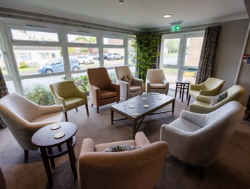 A bright, and vibrant communal lounge area with armchairs around coffee tables by large windows overlooking gardens