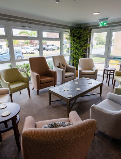 A bright, and vibrant communal lounge area with armchairs around coffee tables by large windows overlooking gardens 