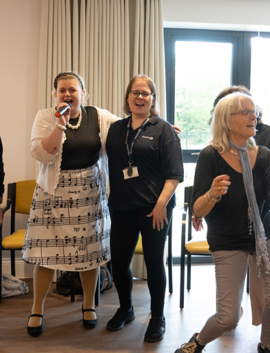 Singer in a musically decorated white skirt, singing while residents dance 