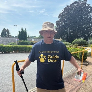 Lee wears a branded Guide Dogs t-shirt and poses with a map and walking stick