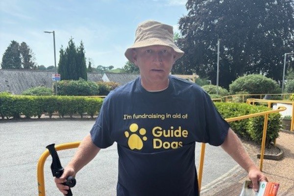 Lee wears a branded Guide Dogs t-shirt and poses with a map and walking stick
