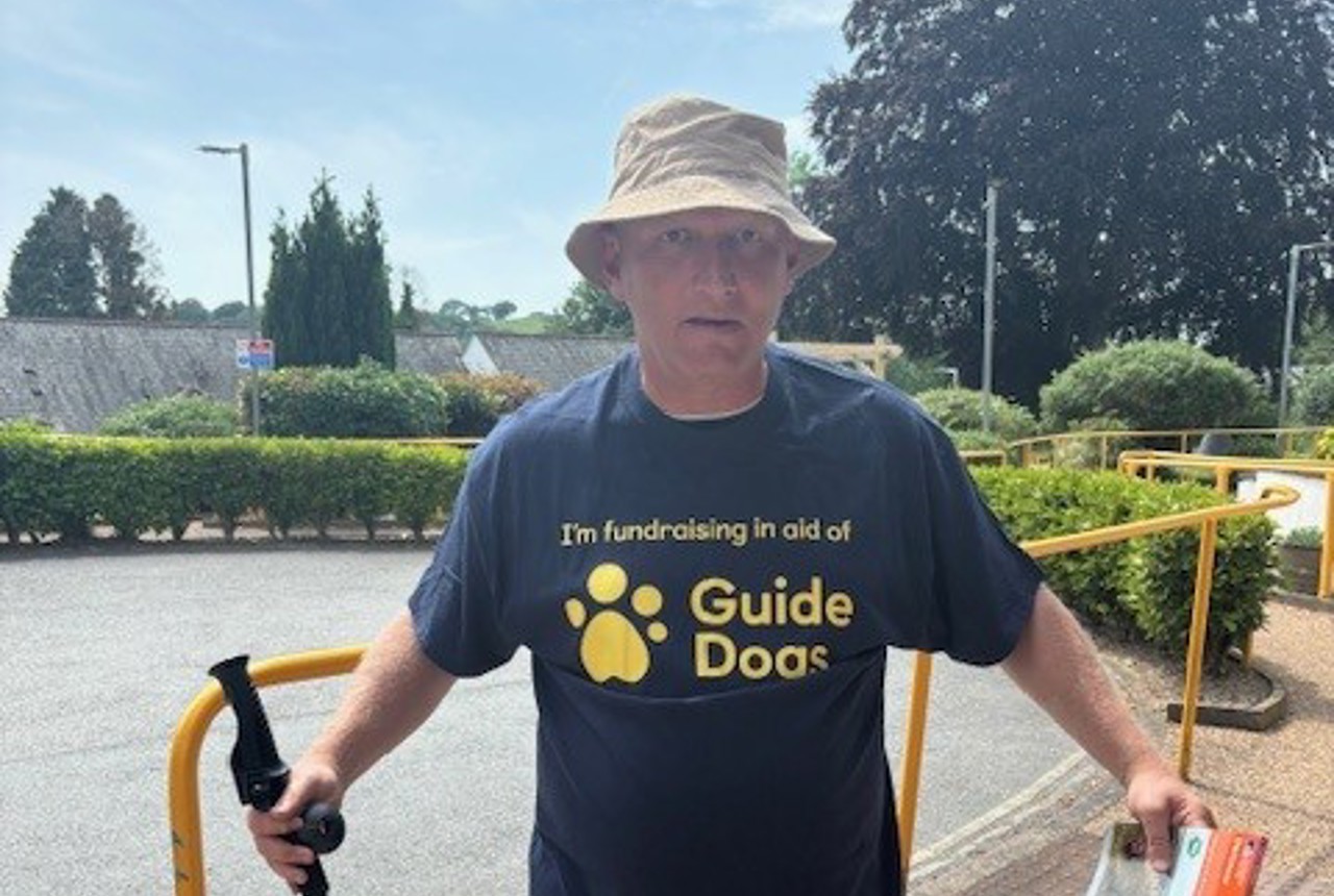 Lee wears a branded Guide Dogs t-shirt and poses with a map and walking stick