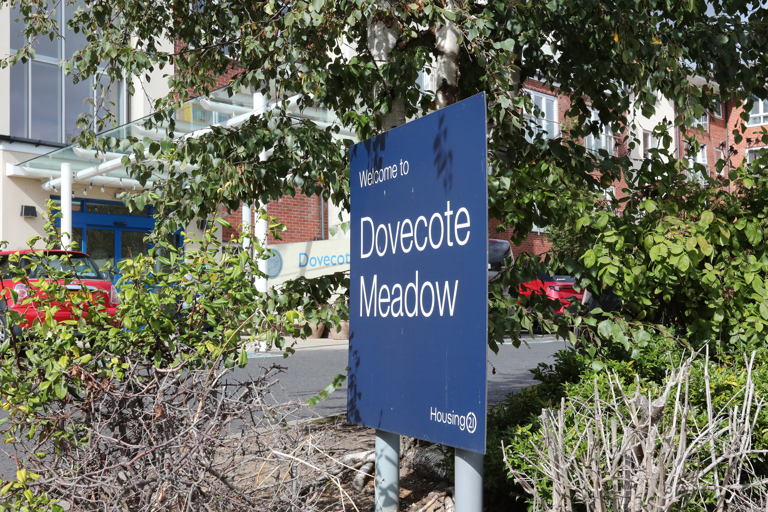 Blue welcome sign for Dovecote Meadow surrounded by trees and planting, with the apartment building and main entrance visible in the background.