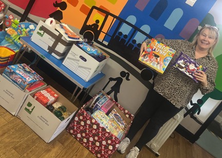 Caron from Ladywood Community Project stands in a room with boxes of donations, holding a board game and a Christmas selection box.
