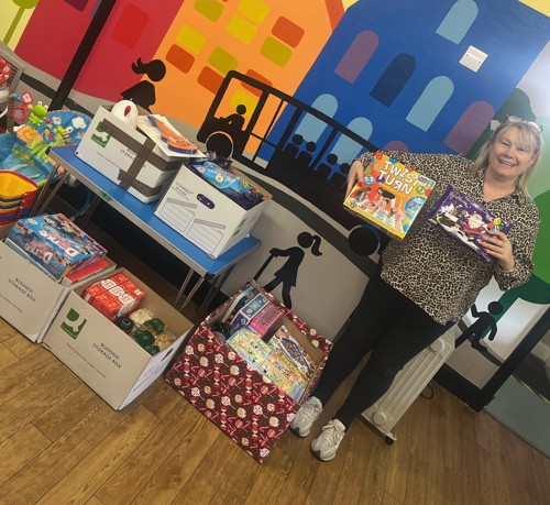 Caron from Ladywood Community Project stands in a room with boxes of donations, holding a board game and a Christmas selection box.
