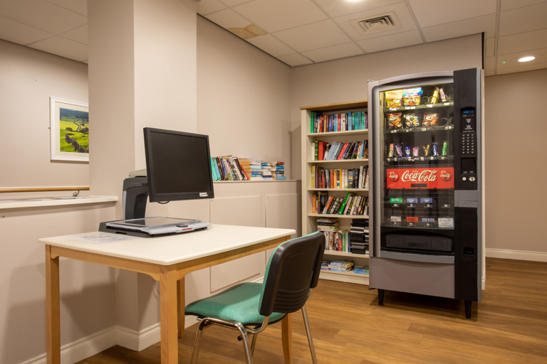 Communal area with a computer workstation, bookshelf stocked with books, and a vending machine offering snacks and drinks.