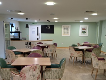 A communal dining area, dining chairs with floral designs pulled up at wooden square tables.