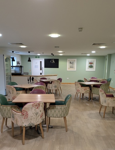 A communal dining area, dining chairs with floral designs pulled up at wooden square tables. 