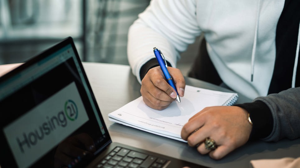 An employee writes in a notebook next to a laptop displaying the Housing 21 logo
