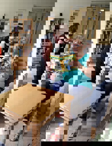 a woman sat down in a chair holding a magazine open and showing a woman standing over her shoulder the contents 