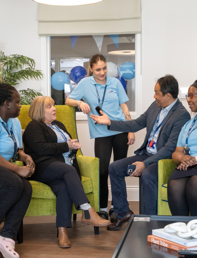 Group of five colleagues in blue branded uniforms talking while sat on green sofas or arm chairs 