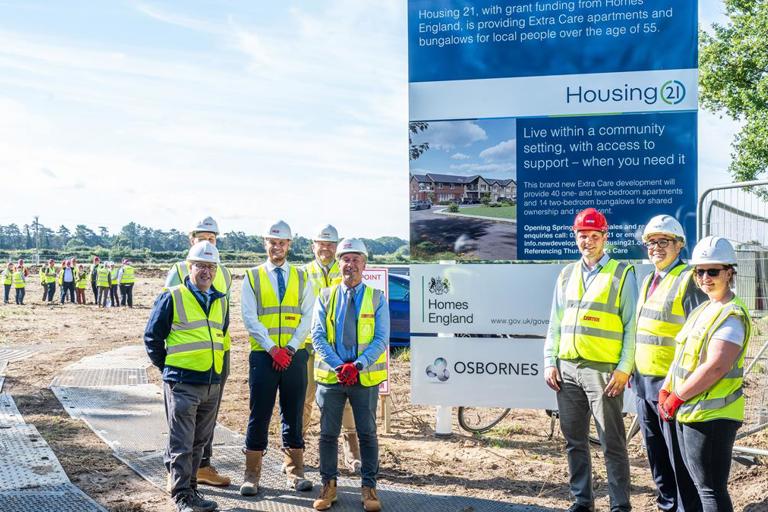Employees of Housing 21 along with special guests wearing high-vis jackets and hard hats standing outside by the sign advertising the new development.