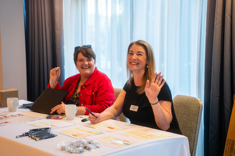 Two ladies sitting at a registration desk smiling and waving
