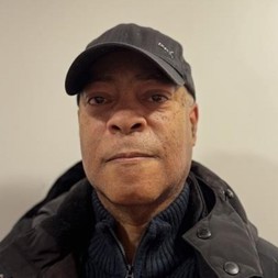 A man wearing a baseball cap, jumper and jacket smiling into the camera