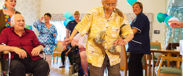 Residents and Care Workers dancing in a communal lounge with chairs and balloons around the room
