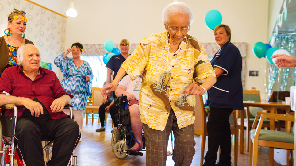 Residents and Care Workers dancing in a communal lounge with chairs and balloons around the room