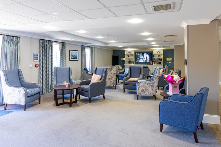 Inside the communal lounge of Oakwood Gardens with light grey carpet and blue arm chairs 