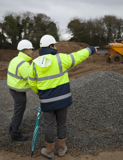 Representatives stood outside on mud and gravel, with a closed umbrella, wearing high vis jackets and hard hats and pointing into the distance 