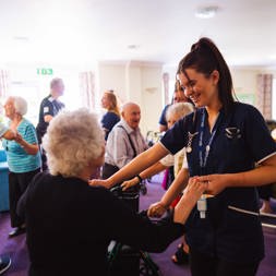 A care worker dancing with a resident in a communal lounge and other residents dancing behind them