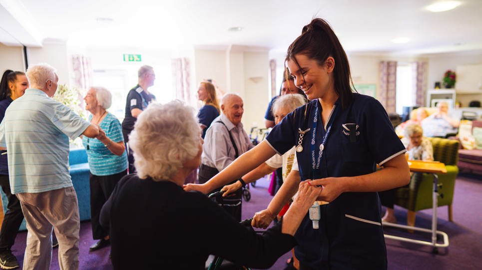 A care worker dancing with a resident in a communal lounge and other residents dancing behind them