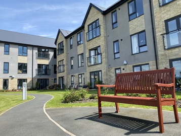 Exterior image of Castlestead View featuring a close of up of a bench in the gardens which are overlooked by a three-storey building