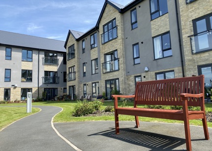 Exterior image of Castlestead View featuring a close of up of a bench in the gardens which are overlooked by a three-storey building