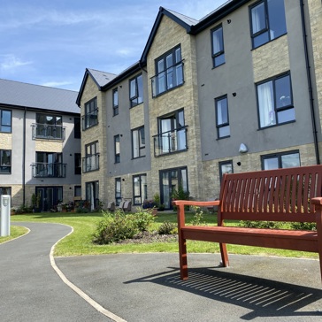 Exterior image of Castlestead View featuring a close of up of a bench in the gardens which are overlooked by a three-storey building