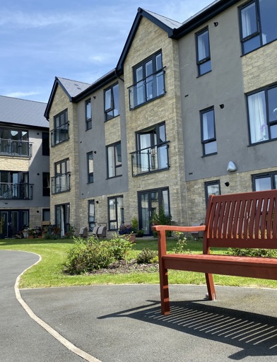 Exterior image of Castlestead View featuring a close of up of a bench in the gardens which are overlooked by a three-storey building 