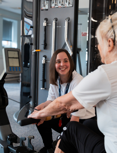 A resident using the rowing machine in the on-site gym with help from an employee 