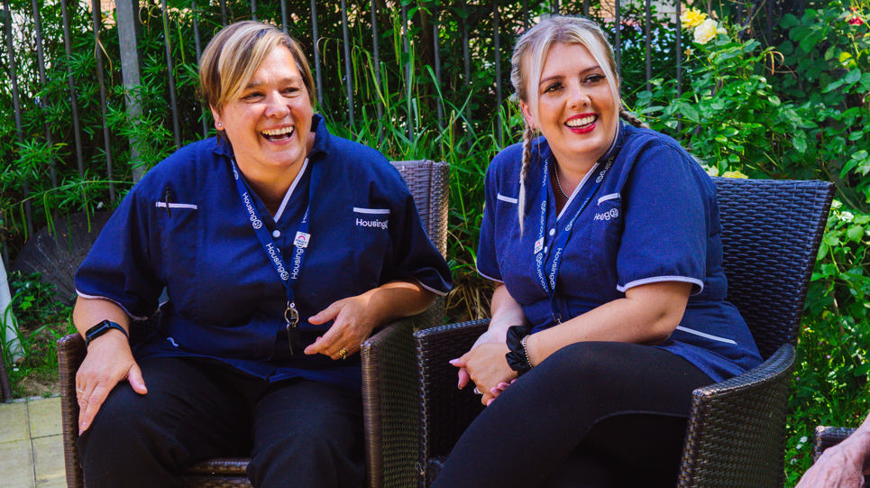 Two Care Workers wearing blue uniforms sat together in communal garden smiling and laughing