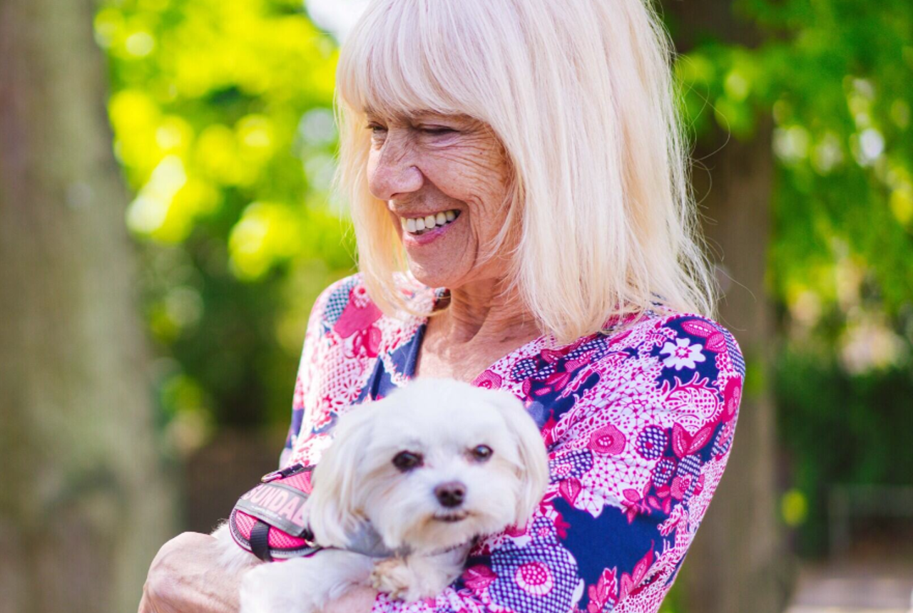 White haired lady in a pink and purple top carrying a small white fluffy dog