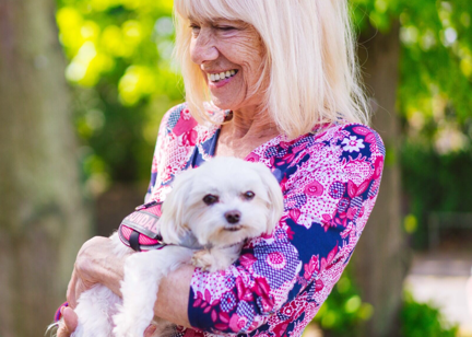 White haired lady in a pink and purple top carrying a small white fluffy dog