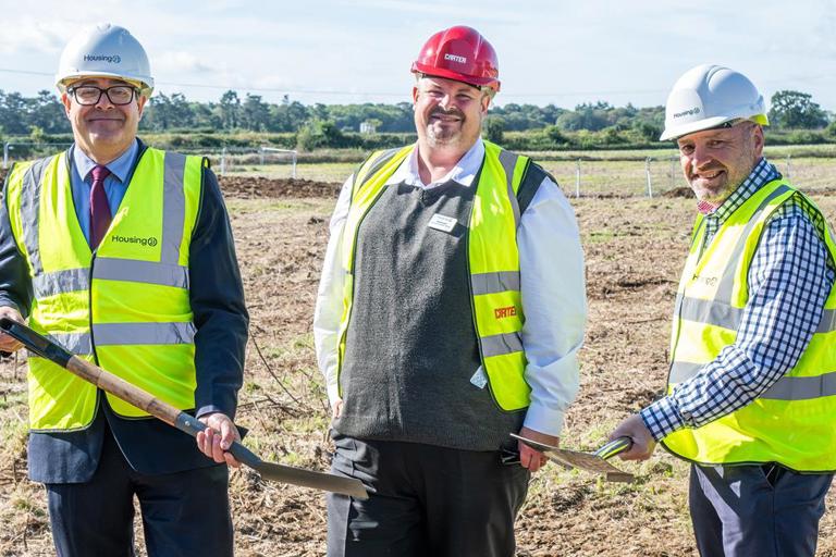 Employees of Housing 21 wearing high-vis jackets and hard hats whilst holding spades standing outside.